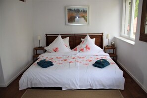 Una cama bien tendida con ropa blanca y motivos florales en rosa, dos mesitas de noche con lámparas, y un cuadro en la pared.