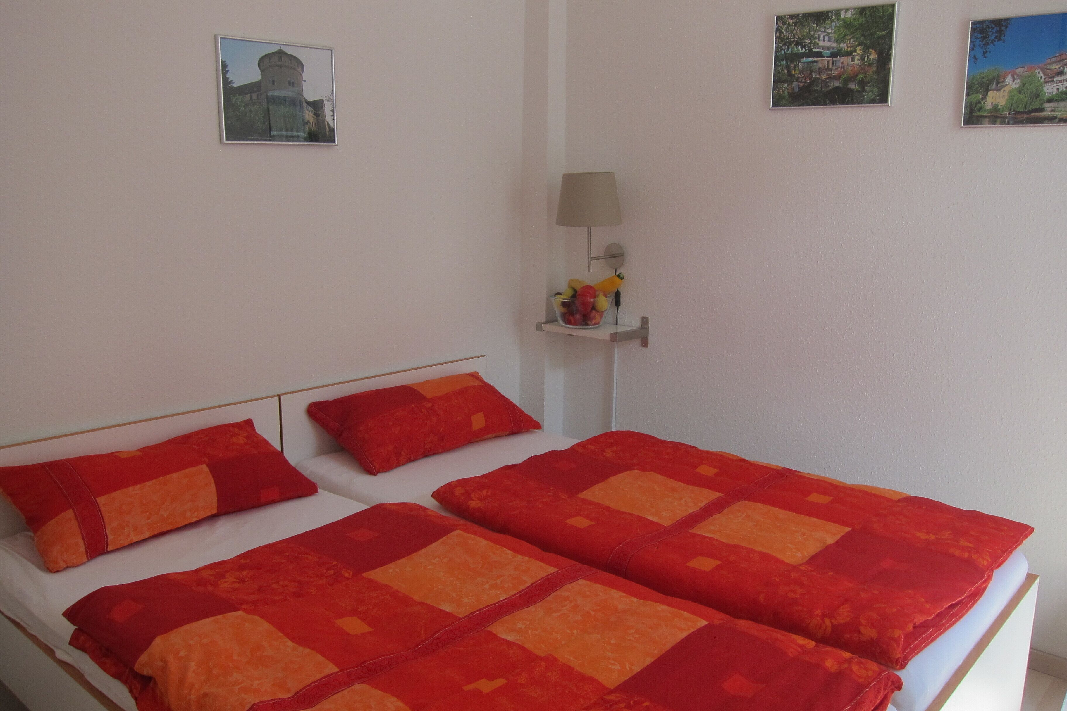 Een tweepersoonsbed met rode en oranje beddengoed, een nachtkastje met een lamp, en twee ingelijste foto's aan de muur.