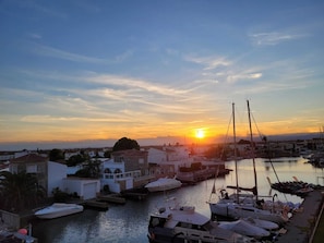 Un puerto deportivo con barcos amarrados, un atardecer de fondo y edificios en primer plano.