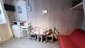 Una habitación compacta con kitchenette, una mesa de comedor con mantel floreado, una silla roja, un espejo redondo pequeño y un reloj de pared.