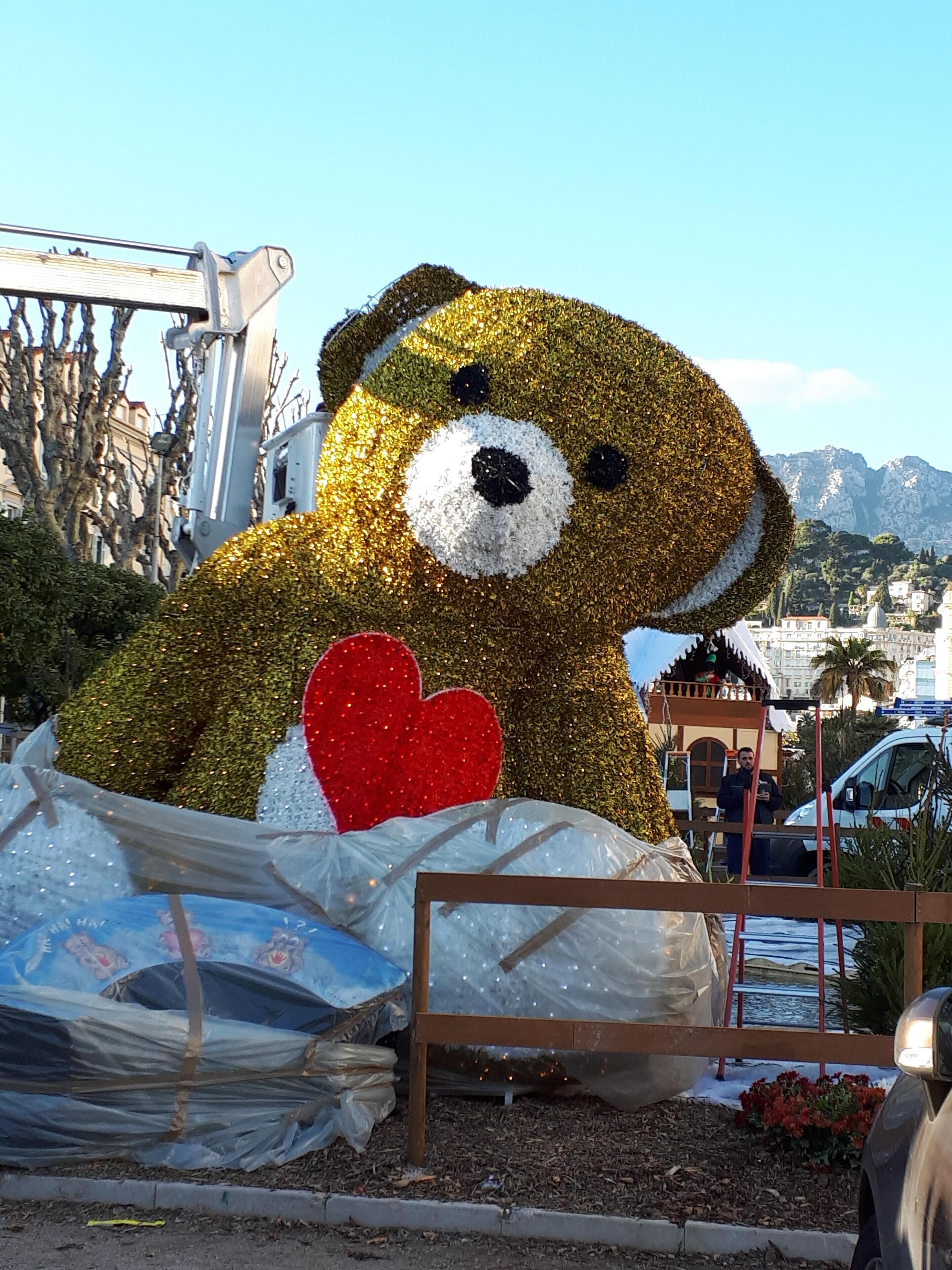 A large, glittery bear sculpture with a heart, surrounded by construction materials and a mountainous backdrop.