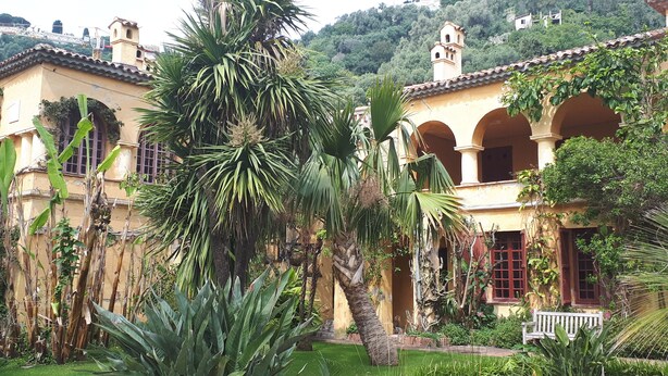 A courtyard with a palm tree, a bench, and a building with arches and a tower. - --- Beachfront Studio ---- (Menton)