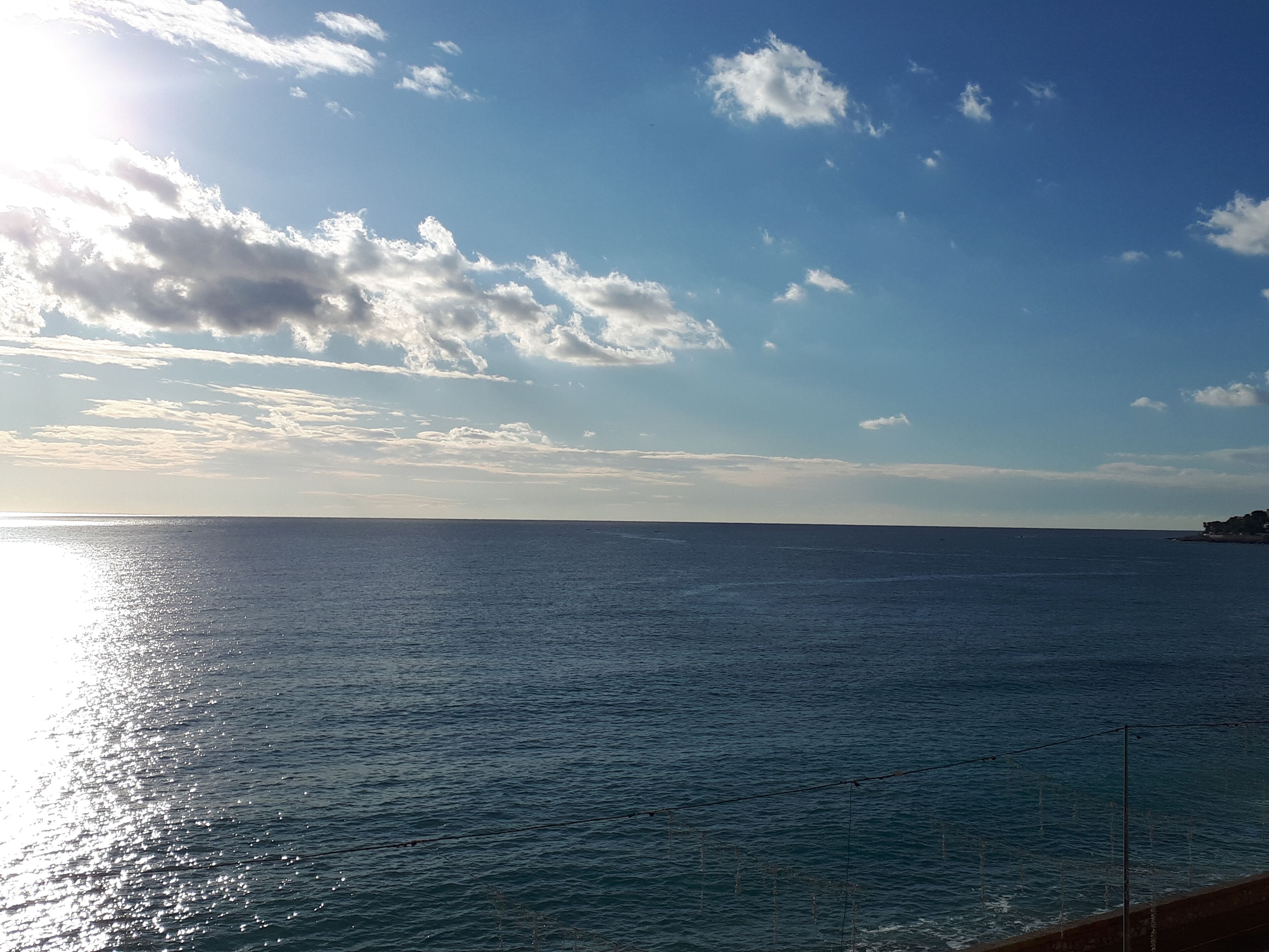Vista costanera amb cel clar i mar tranquil·la, una línia de costa llunyana i una línia elèctrica sobre l'aigua.
