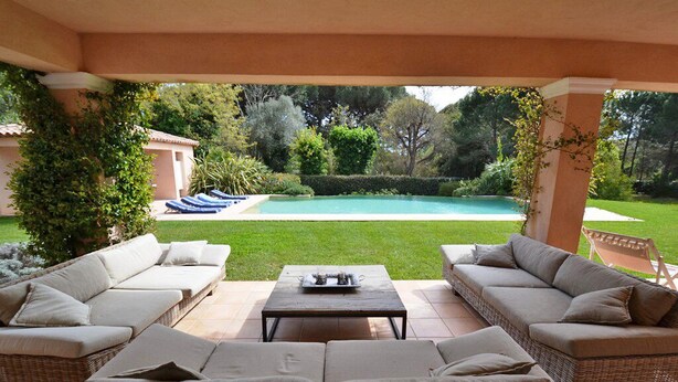 Un'area salotto esterna coperta con divani in vimini e un tavolino da caffè, con vista su una piscina e vegetazione lussureggiante.