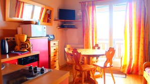 A compact kitchen with a microwave, coffee maker, and a small TV on a shelf. There is a dining table with chairs and a window with curtains.