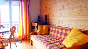 A cozy room with a wooden floor, a colorful checkered sofa with yellow cushions, a wooden side table, and a window with curtains.