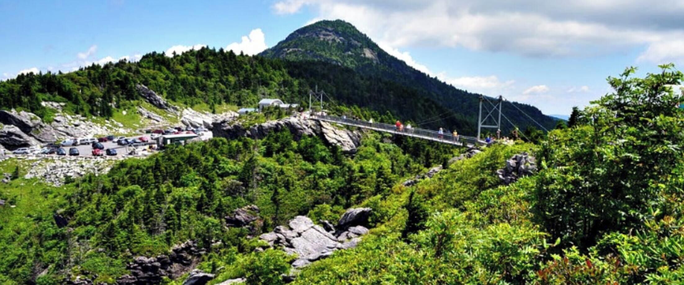 A mountainous landscape with a suspension bridge, dense greenery, and a parking area.