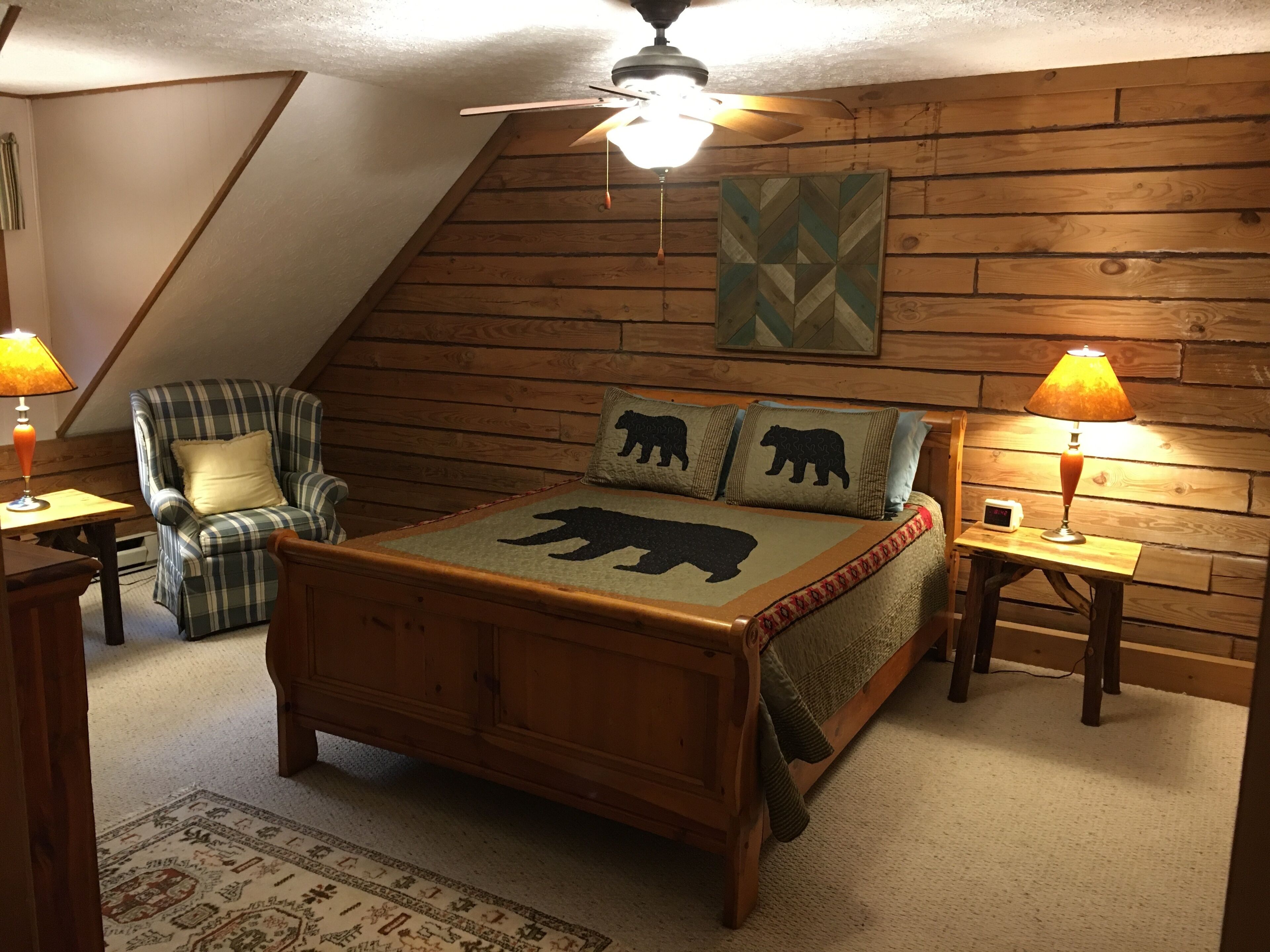 Een gezellige slaapkamer met een schuine kap, een houten bed met een sprei met berenprint, een ruitjes fauteuil en twee bedlampjes.