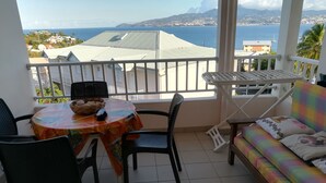 A balcony with a table and chairs, a colorful sofa, and a view of the sea and buildings. - STUDIO ANSE MITAN TROIS ILETS MARTINIQUE (Trois Ilets)