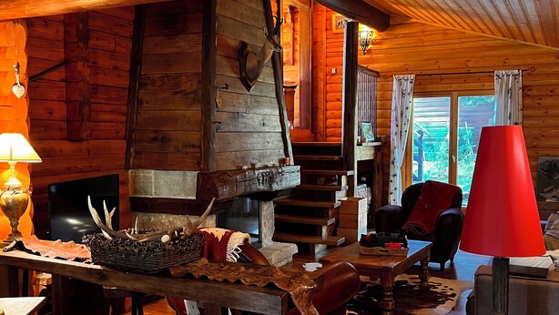 A rustic cabin interior with wooden walls, a stone fireplace, a wooden staircase, and a dining area with a red lamp.