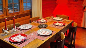 A wooden dining room with a table set for four, featuring red and white tableware, candles, and a view of the outdoors through large windows.