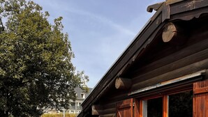 A wooden deck with a picnic table and chairs, a wooden cabin with large windows, and a view of a tree and hillside.