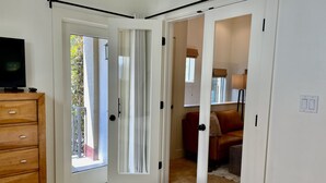 Une pièce comprenant une commode en bois, un lit et une porte donnant sur une autre pièce où se trouvent un canapé rouge et une lampe.