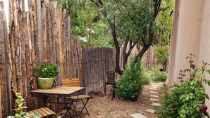 Une table et des chaises en bois sont placées devant une clôture en bois rustique, et l’ensemble est entouré d’un chemin de gravier et de diverses plantes.