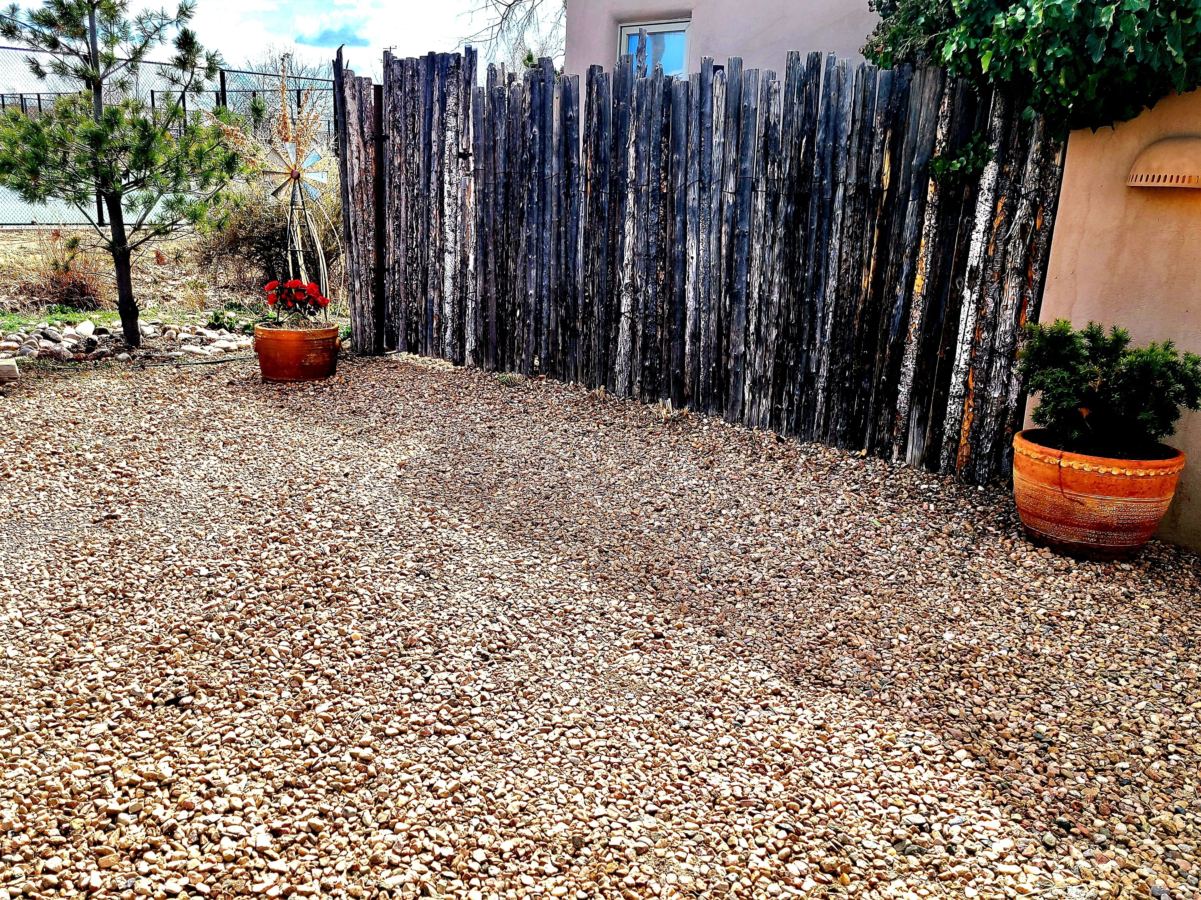 A gravel patio with a wooden fence, a potted plant, and a building in the background.