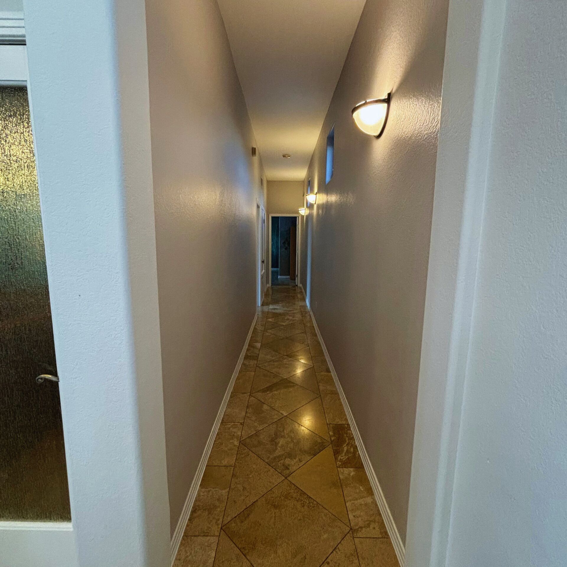A hallway with white walls, a tiled floor, and wall-mounted lights.