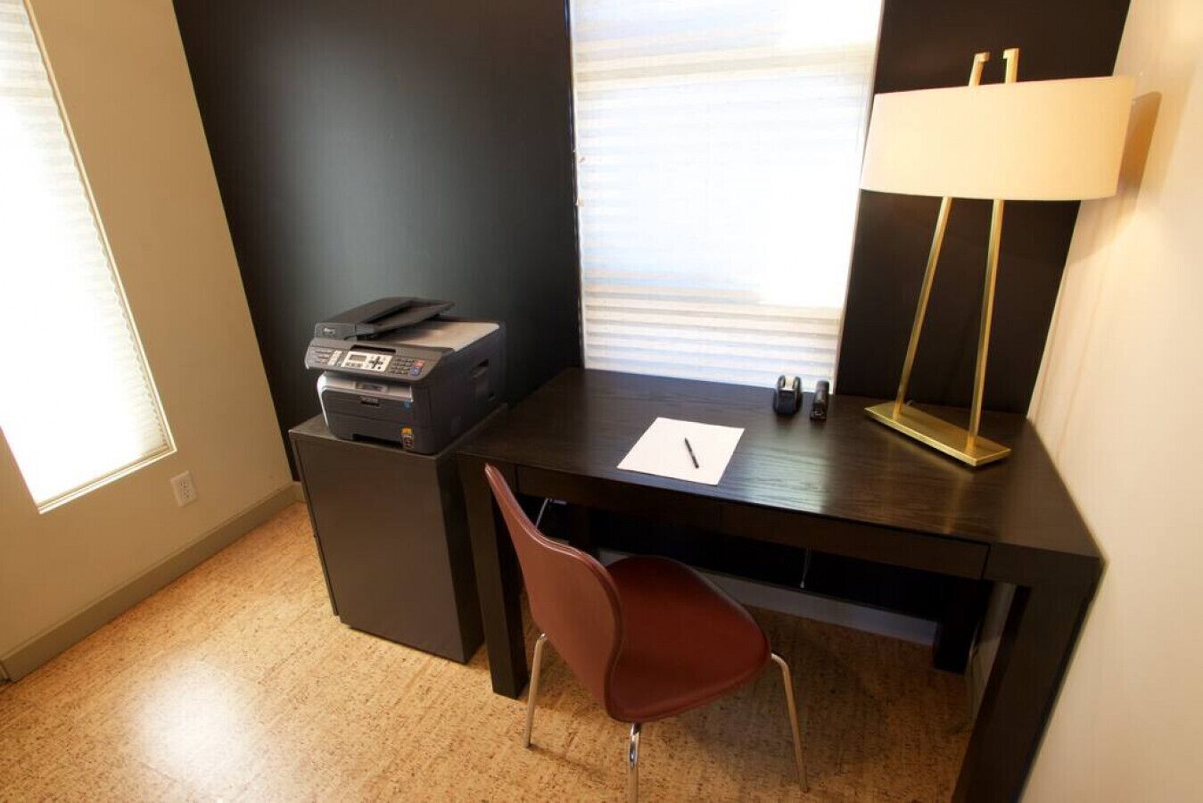 Un pequeño espacio de oficina con un escritorio, una silla, una impresora, una lámpara y una ventana.