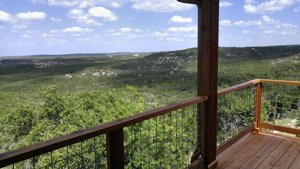 A wooden deck with a metal railing overlooking a forested landscape with hills and scattered buildings. - VIEWS! Texas Hill Country - Terra Vista (Dripping Springs)