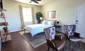 A bedroom with a bed, two leather chairs, a nightstand, a lamp, a potted plant, and a ceiling fan.