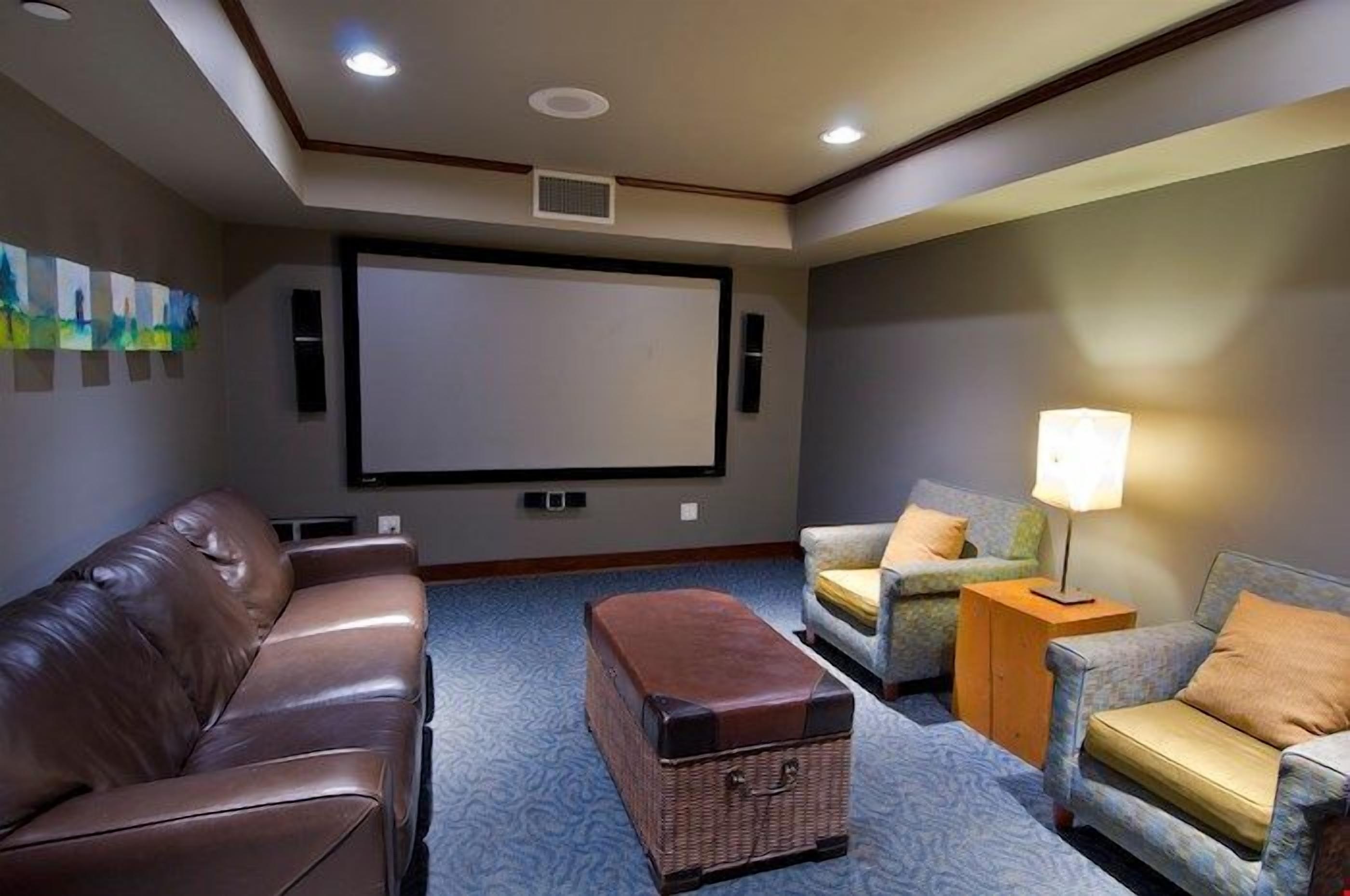 Una sala de cine en casa con un sofá de cuero marrón, dos sillones, una mesita con una lámpara y una pantalla grande.