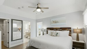 Quarto com uma cama grande, um ventilador de teto, uma mesa de cabeceira com abajur e vista para o banheiro através de uma porta aberta.