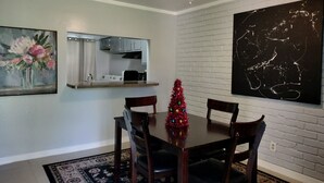 Un área de comedor con una mesa de madera, cuatro sillas, un pequeño árbol de Navidad, un ventilador de techo, una cocina con estufa y armarios, y un cuadro abstracto de gran tamaño en la pared.