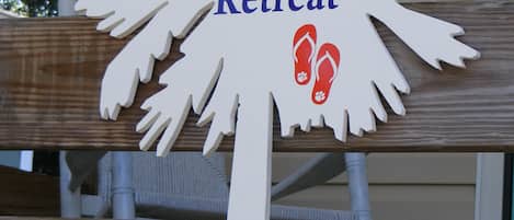 En träskylt med texten ”Sandy Feer Retreat” och en silhuett av en palm, en halvmåne och ett par flip-flops.