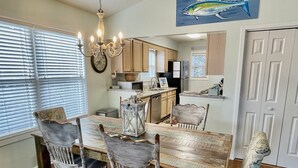 A dining area with a wooden table and four chairs, a chandelier, a kitchen in the background, and a fish painting on the wall. - Sandy Feet Retreat-1/2 Block to Beach, Pet Friendly, Wi-Fi & Optional GC Rental  (Surfside Beach)