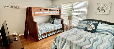 A bedroom with a bunk bed, a single bed, a ceiling fan, a bedside table with a lamp, and a wall sign that reads "BEACH CLUB".