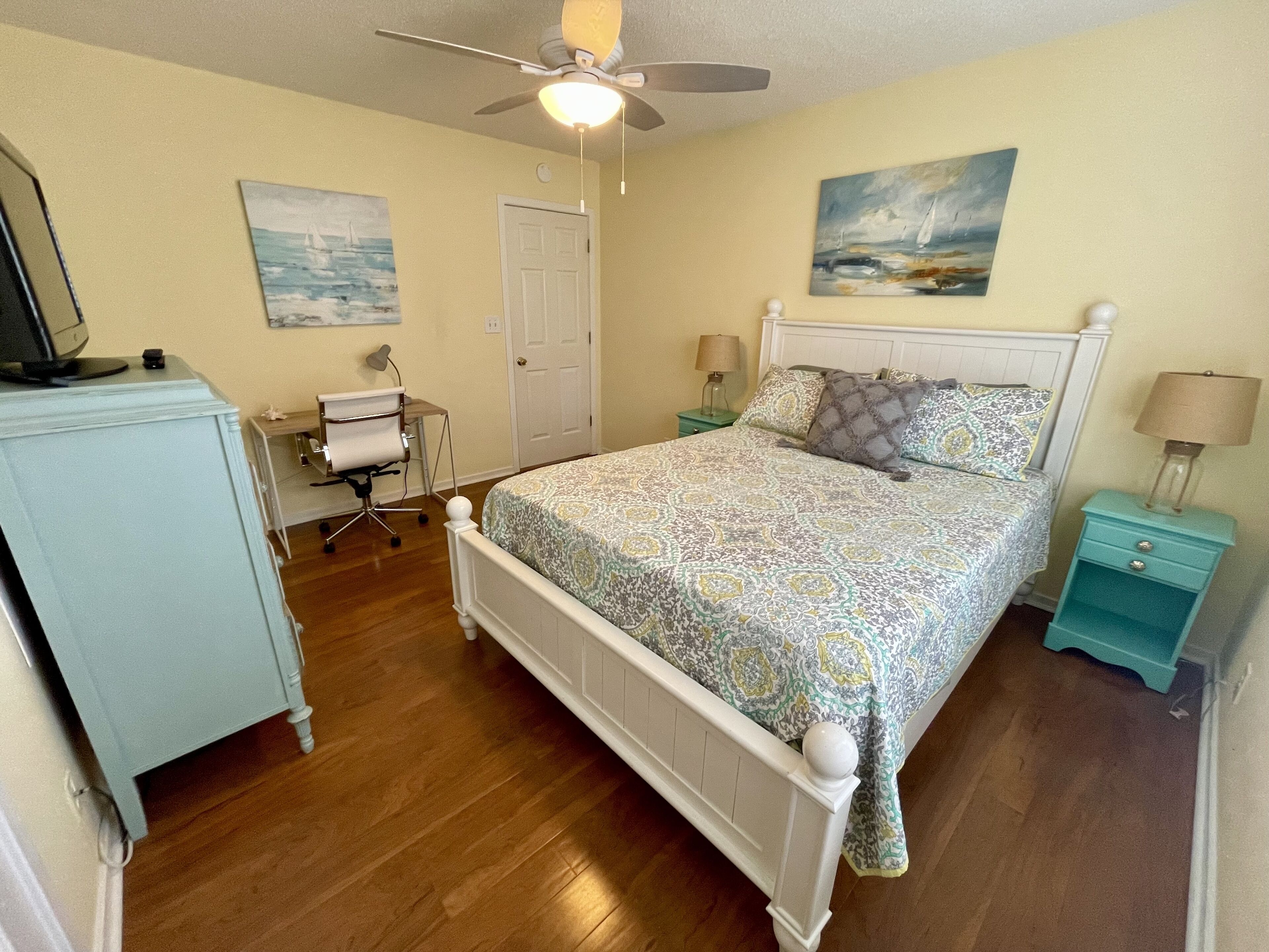 A bedroom with a bed, a desk, a chair, a ceiling fan, and two paintings on the walls.