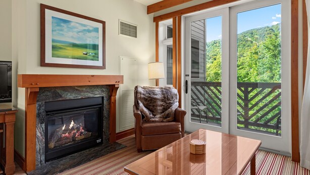 A room with a fireplace, a framed landscape painting, a chair, a table, and a balcony with a view of greenery. - Luxury Studio Ski in -ski out Mt Mansfield Views, Mountain Golf Course Access (Stowe)