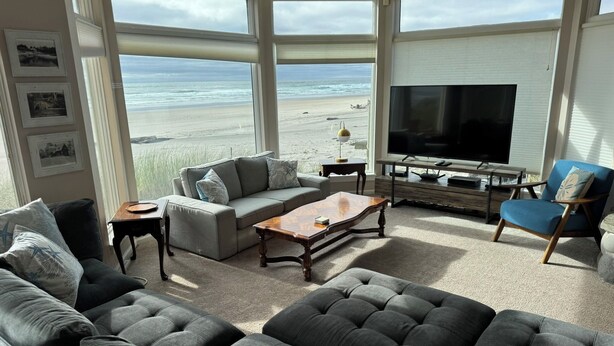 A spacious living room with a large window overlooking a beach, a television, and comfortable seating.