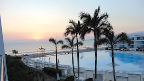 A resort with a large pool, palm trees, and a view of the ocean at sunset.