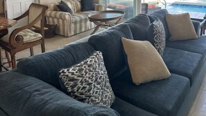A spacious living area with a large gray sofa, striped armchairs, and a small round table. A large window offers a view of the ocean and a balcony with a railing. - Relax in Paradise on St. Croix's North Shore - Cane Bay! (Christiansted)
