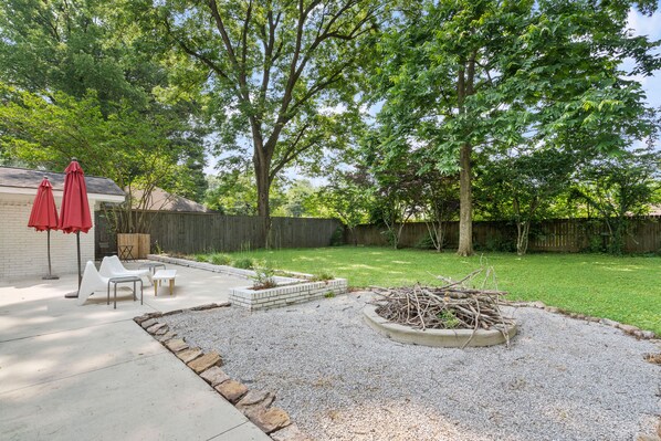 Relax in the serene backyard patio surrounded by a lush lawn and trees, featuring a fire pit and comfortable seating.