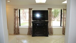A room with a flat-screen TV on a black stand, large windows with sheer curtains, and a tiled floor. - Wonderful Whispering Hills (Tucson)