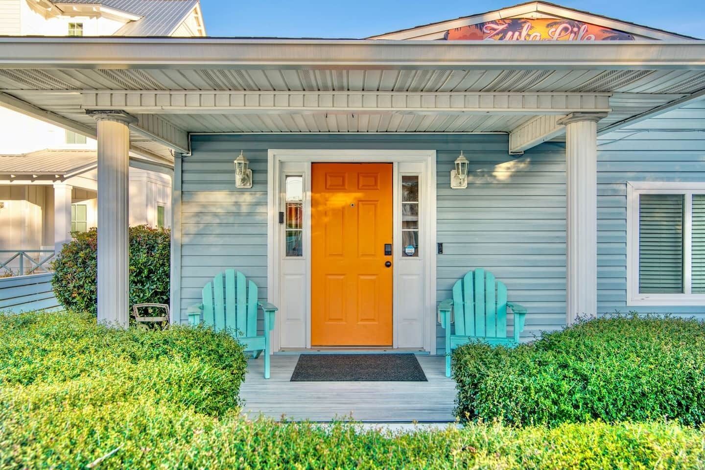 Uma casa com uma porta frontal laranja vibrante, duas cadeiras Adirondack azul claro e uma varanda coberta com colunas.