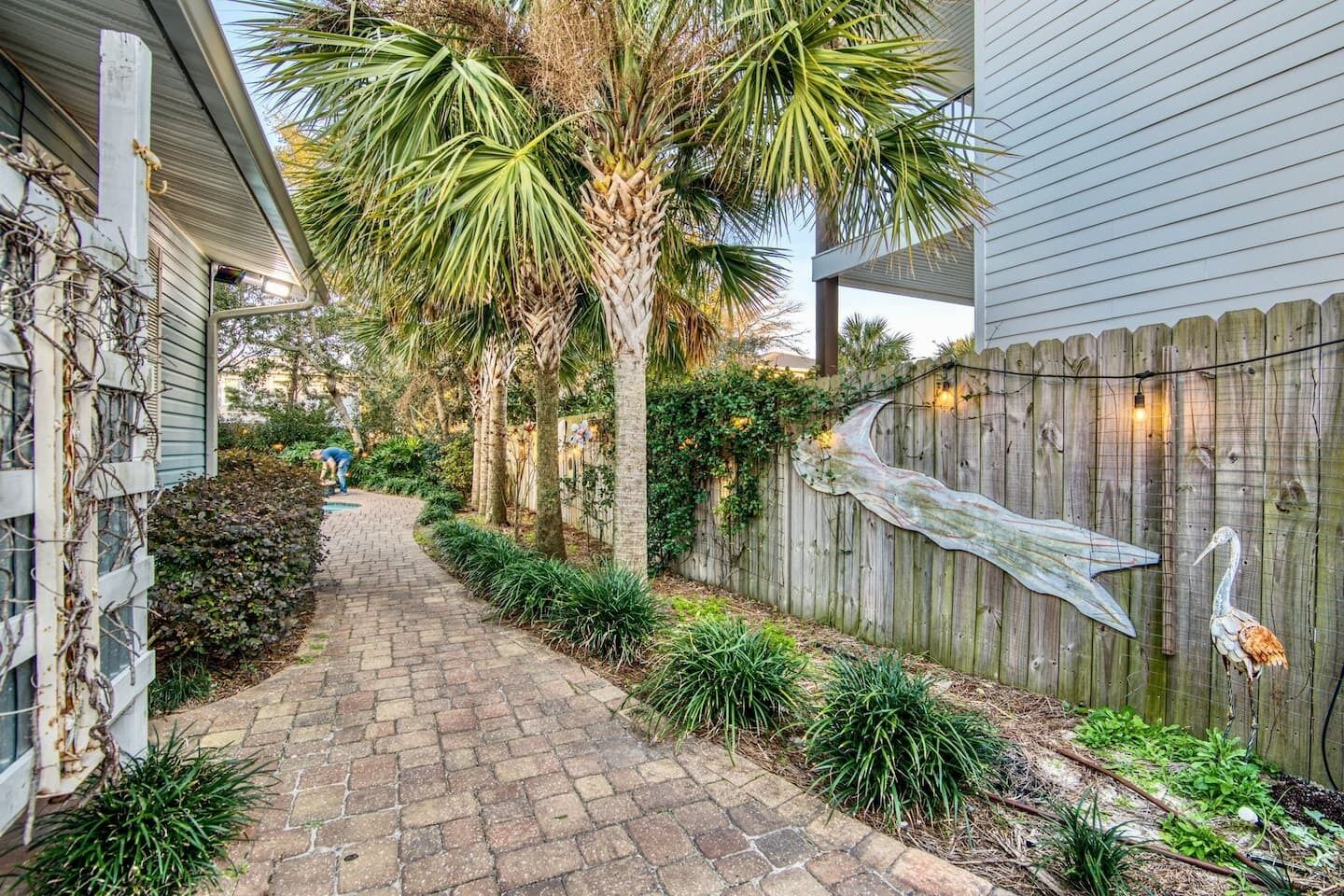 A narrow brick pathway flanked by a wooden fence and palm trees, leading to a house with a porch.