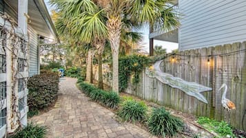A narrow brick pathway flanked by a wooden fence and palm trees, leading to a house with a porch.