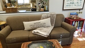Een woonkamer met een bruine bank, een decoratief kussen, een salontafel met een glazen blad en een bloemistisch ontwerp, en een boekenkast met boeken en een ingelijst kunstwerk.
