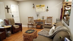 Een gezellige woonkamer met een bruine leren bank, een houten bijzettafeltje en een rieten mand. Er staat een houten bureau met een stoel, een kruis aan de muur en een bordje met de tekst 'Gallo Wines Sonoma'.