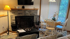 Un salon doté d’une cheminée en pierre, d’un téléviseur à écran plat, d’une table à manger avec des chaises et d’une grande fenêtre avec des stores.