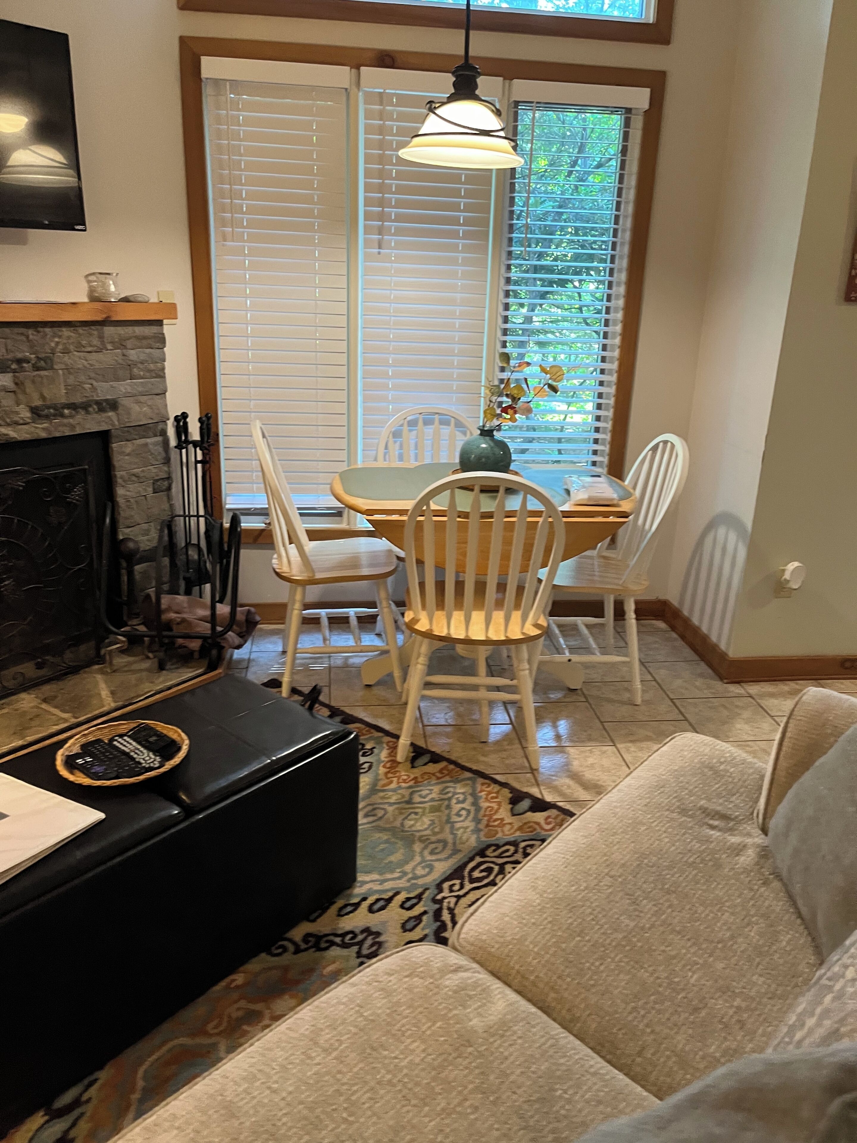 A cozy living room with a fireplace, a dining area with a table and chairs, and a large window with blinds.