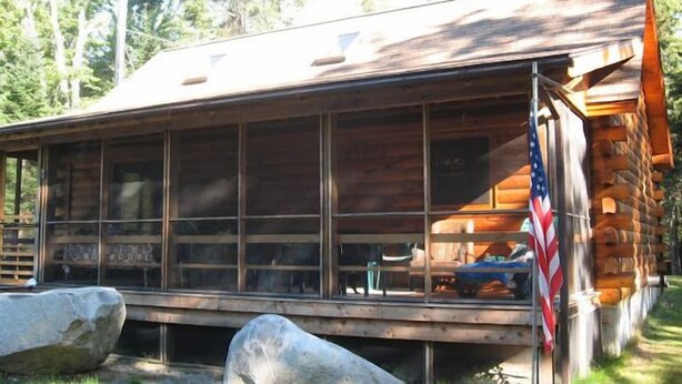A log cabin with a large windowed porch, a flag, and a rock in front. - Hand Crafted Log Cabin (St George)