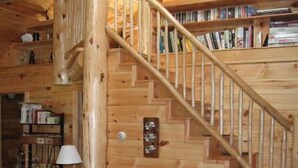 A cozy log cabin interior with a wooden staircase, a leather sofa, and a bookshelf filled with books. - Hand Crafted Log Cabin (St George)