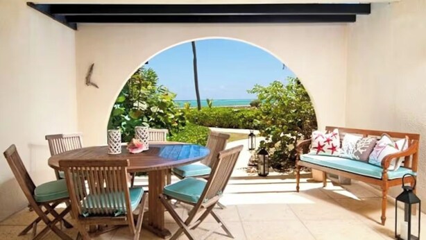Eine überdachte Terrasse mit einem runden Holztisch und Stühlen, einer Couch mit Kissen und Meerblick durch einen Bogen.