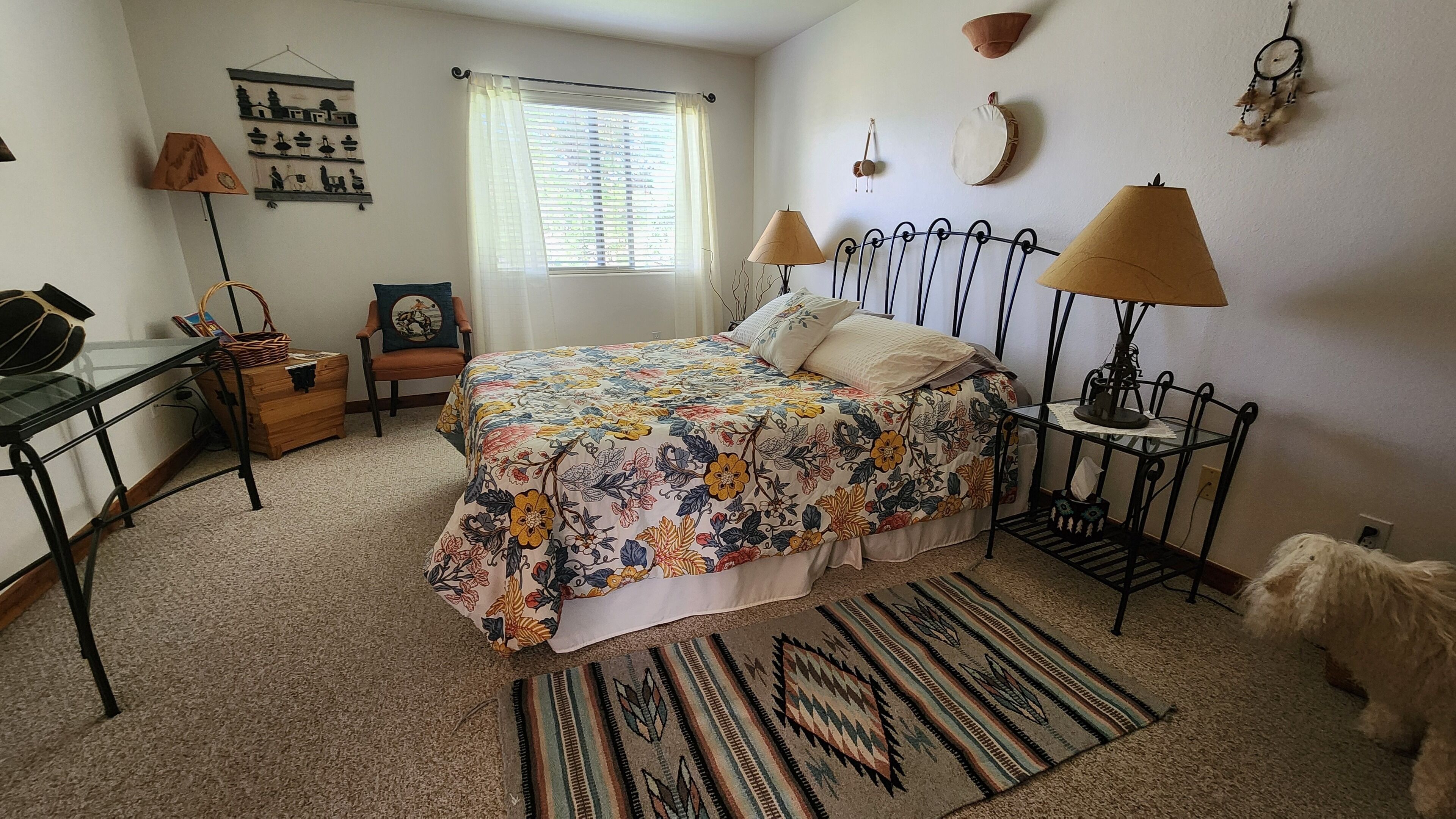 A bedroom with a floral bedspread, a patterned rug, a desk with a lamp, and a small dog.
