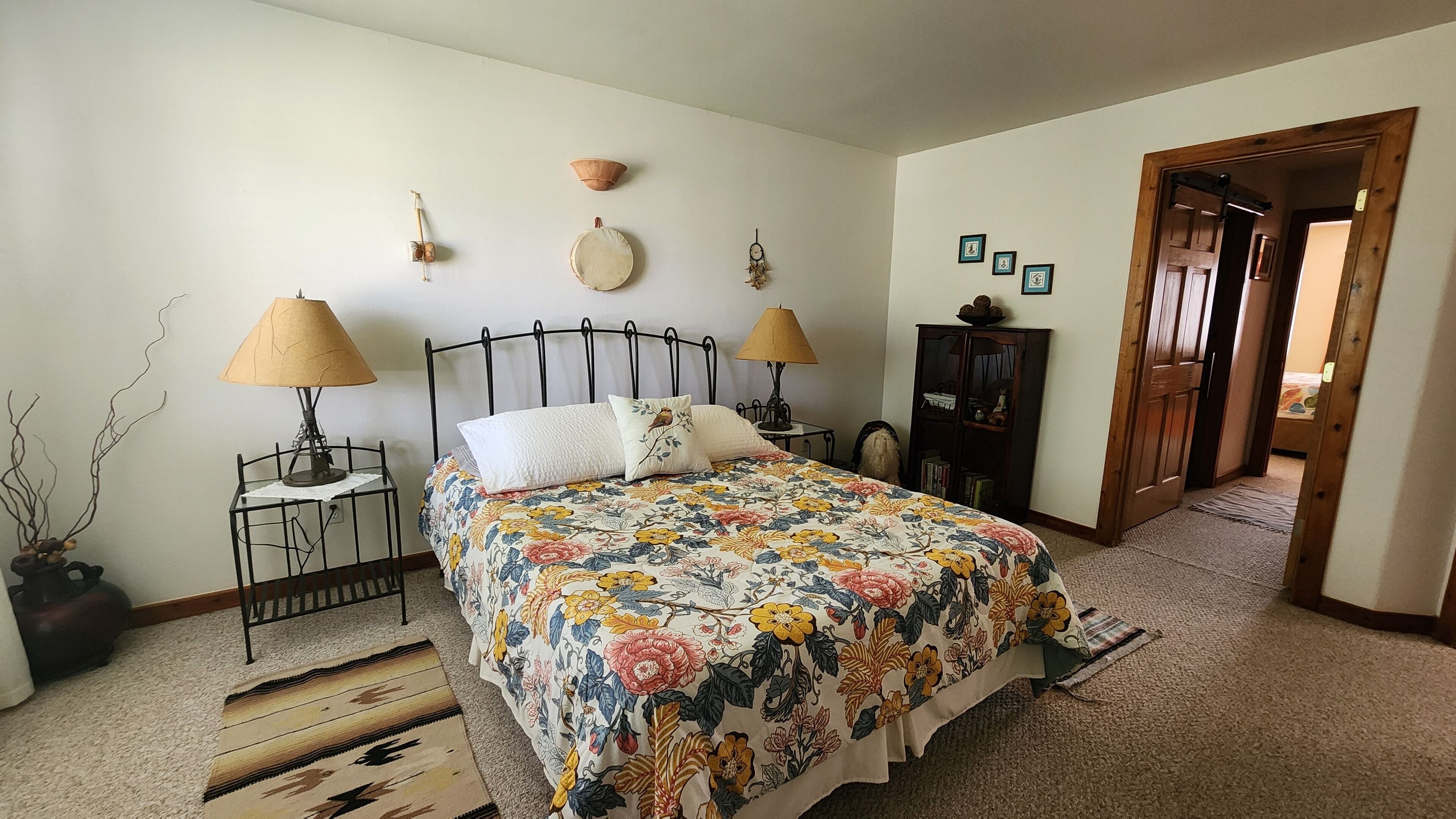 A bedroom with a bed, two bedside tables, a lamp, a dresser, and a door leading to another room.
