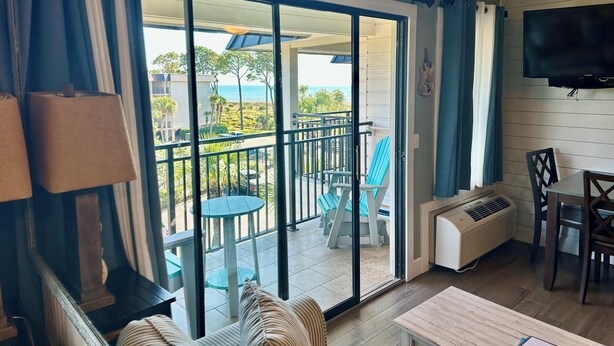 A room with a sliding glass door leading to an outdoor patio with a table and chairs, a ceiling fan, and a flat-screen TV mounted on the wall. - SeaGem-Oceanview! Steps to ocean Many Upgrades New Reno (Hilton Head Island)