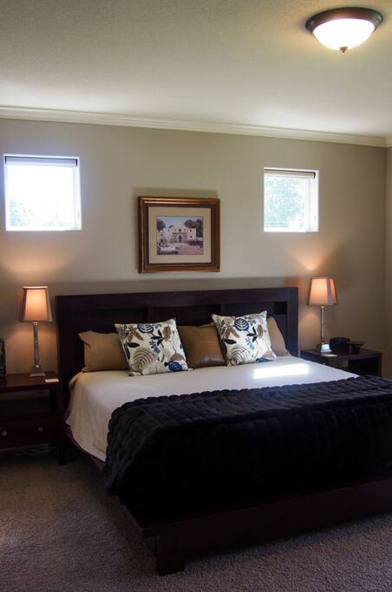 A bedroom with a large bed, two bedside tables, a framed picture on the wall, and two windows.